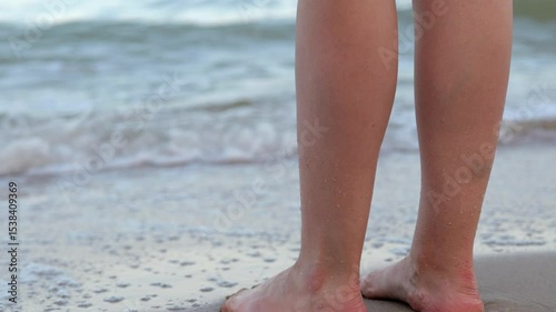 A close-up of bare feet on a sandy beach, with a wave breaking and foaming around them.