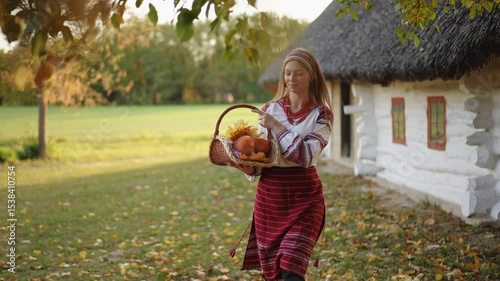 A woman in beautiful traditional attire joyfully holds a basket of pumpkins in a picturesque autumn landscape
