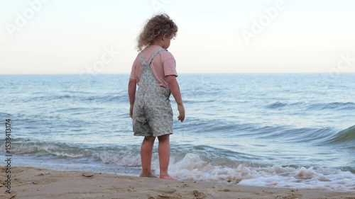A young child with curly hair squats at the edge of the sea, playing in the sand and water.