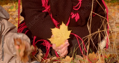 In a scenic grassy area, a woman elegantly poses in her chic brown hat and stylish black sweater, gently holding a colorful autumn leaf and enjoying the beauty of the season