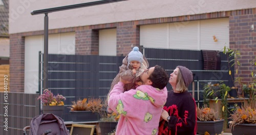 In a cozy area with planters, a man in a pink hoodie lifts a child in a blue hat as a smiling woman observes, highlighting community and affection on a pleasant autumn day