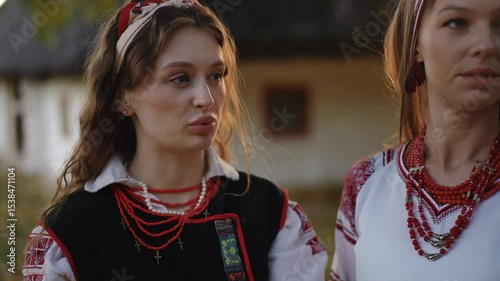 Close-up of two young women in traditional embroidered Ukrainian costumes talking in front of an old thatched house, one gesturing while the other listens attentively.