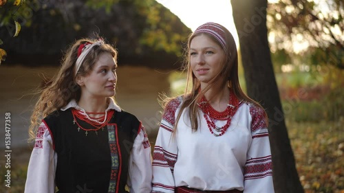 Close-up of two young women in traditional Ukrainian outfits walking quickly but captured in slow motion, with soft sunlight and an old thatched house in the background.