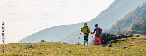 Couple hikers trek up a rocky mountain trail, enjoying a scenic view above the clouds, with majestic peaks in the background.