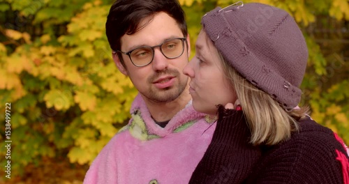 In an autumn park, a couple shares a tender moment. The man in a pink hoodie and the woman in a black sweater and gray hat enjoy a stroll through the vibrant, picturesque landscape