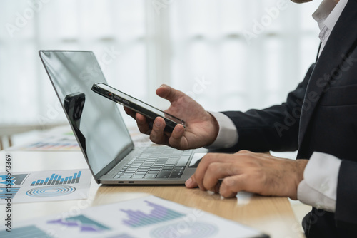 Business professional in formal attire checking smartphone while working on laptop at office desk with financial charts and graphs in the background