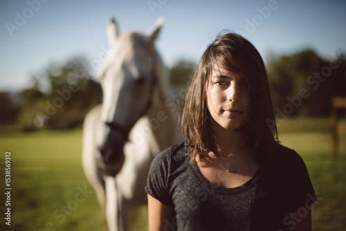 Woman standing in sunlit farm pasture with white horse behind and showing pendant necklace photo