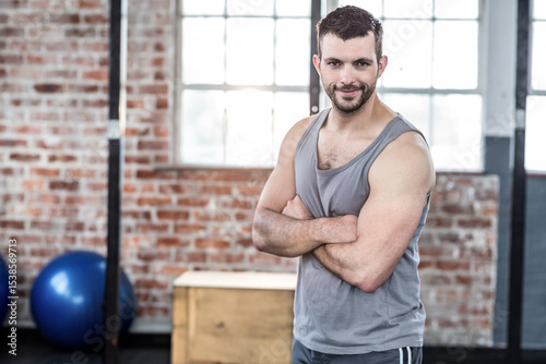 Man standing with crossed arms inside gym by wooden box, metal beam, exercise balls, copy space