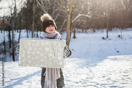 Boy aged seven standing in snowy woodland clearing wearing padded coat and holding silver gift box photo