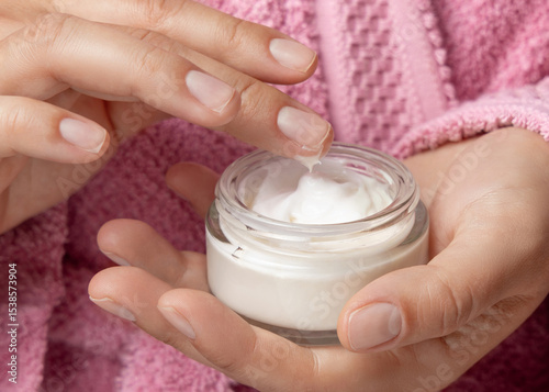 Young woman in pink bathrobe touching cream with finger and holding a cosmetic jar closeup