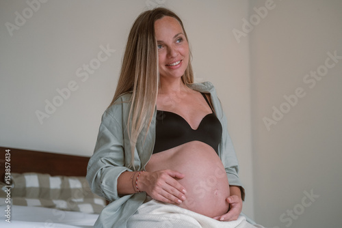Portrait of a pregnant young woman. She is sitting on bed in the bedroom and stroking her round belly in anticipation of arrival of the baby. Third trimester of pregnancy, medical care and childbirth