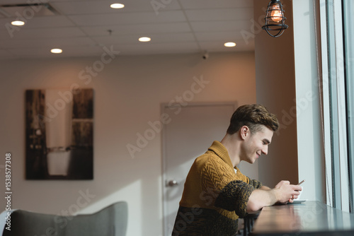 Man sitting on ledge by windows smiling at smartphone wearing sweater beside cage light, copy space