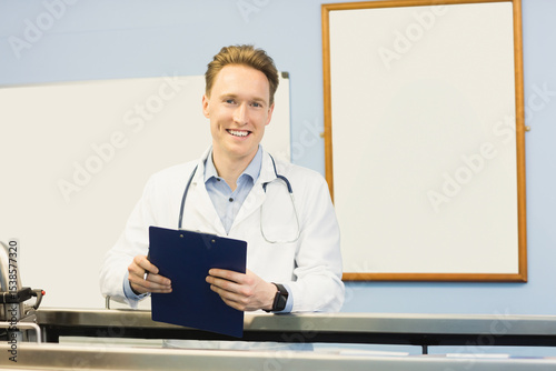 Male doctor leaning on reception desk wearing stethoscope and holding clipboard while smiling