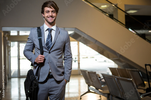 Man standing and smiling at camera in airport terminal near escalator, carrying black shoulder bag photo
