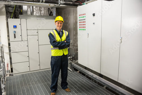 Male technician wearing hard hat standing in equipment room by control panel and indicator lights