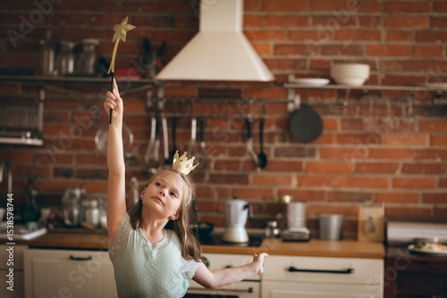 Girl posing wearing gold toy crown and holding star-tipped magic wand above head in rustic kitchen
