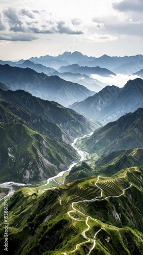 Serpentine road winding through verdant mountain terraces and valley landscape beneath a hazy sky, distant peaks fading into the background.