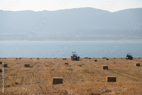 Hay bales have been gathered in the field and two tractors are working in the field. In the background there is a large lake and mountains. photo