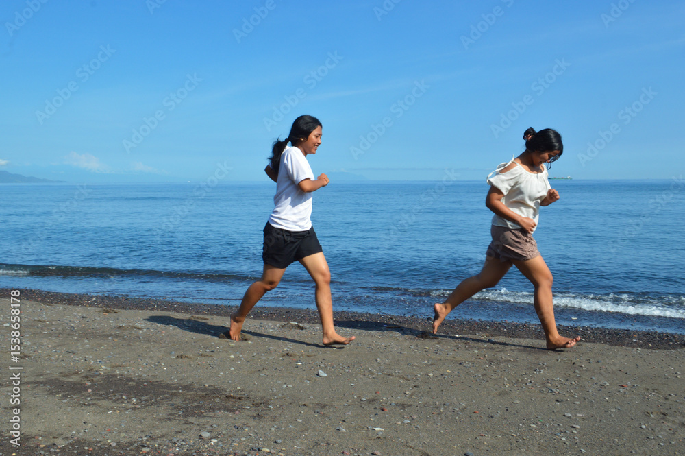 Fototapeta premium Two teenage girls run freely along the sandy beach, letting the ocean breeze guide them as they soak up the peaceful yet lively atmosphere of the bright morning waves.