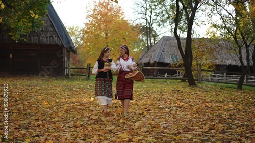 Two girls in traditional embroidered clothing walk barefoot across a carpet of fallen leaves, carrying a basket and talking near old wooden village houses.