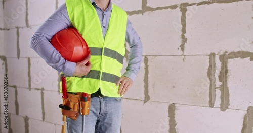 Builder takes off helmet after successfully completing work. Specialist with tool belt stands near large cinder block wall in apartment hall at renovation