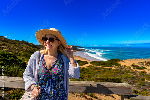 Happy mature woman in summer hat tourist standing  at viewpoint to Monte Clerigo beach on Algarve coast on sunny spring day. Front view