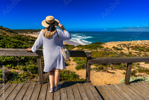 Mature woman in summer hat tourist standing at viewpoint to Monte Clerigo beach on Algarve coast on sunny spring day. Back view