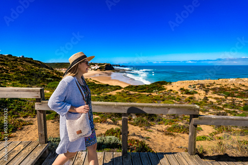 Mature woman in summer hat tourist walking on wooden stairs overlooking at Monte Clerigo beach on Algarve coast on sunny spring day. Side view