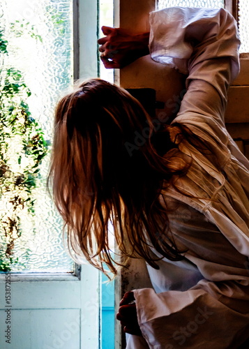 A person in a white garment leans against a window frame, their face obscured by their hair.