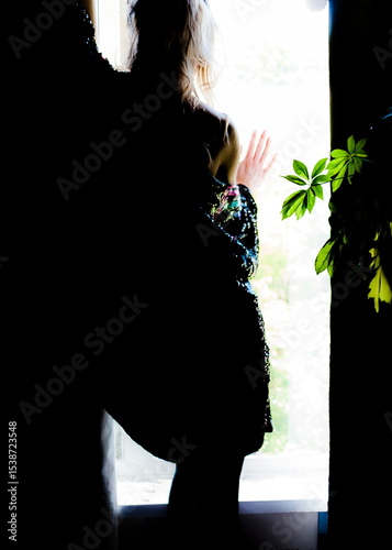 A woman in a sequined dress stands by a window, her hand touching the glass.