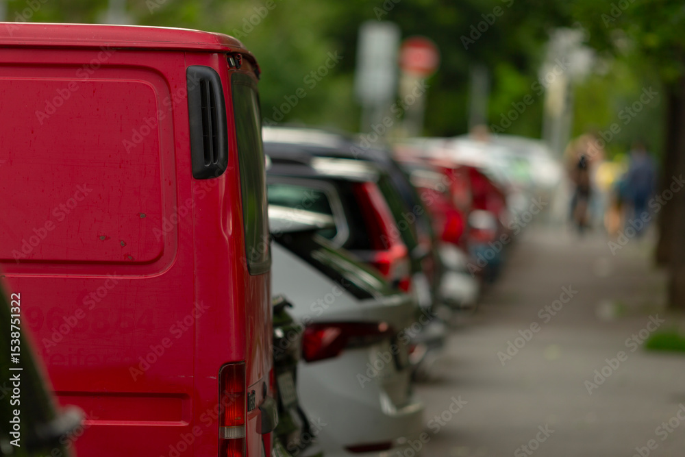 Fototapeta premium Along a bustling city street, a long row of parked cars, showcasing a variety of automobiles, includes a particularly striking red van that captures the attention of many passersby nearby