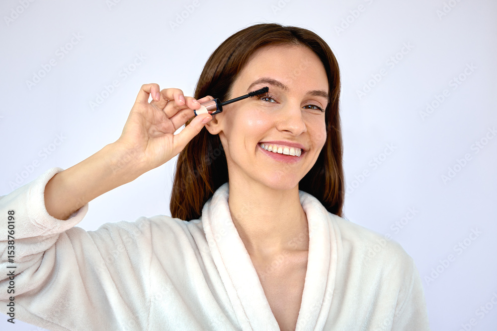 Beautiful young happy brunette dyeing eyelashes with mascara in a white bathrobe on the white background Beautiful young happy brunette dyeing eyelashes with mascara in a white bathrobe on the white background