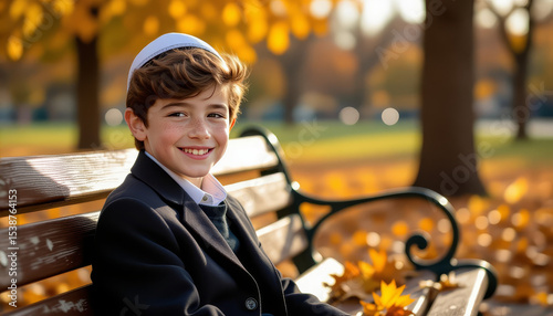 Smiling orthodox Jewish boy with kippah sitting on bench in autumn park
