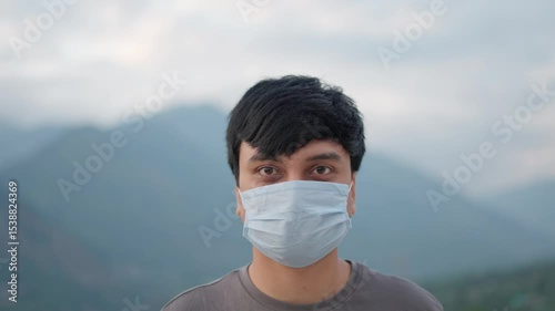 4K Portrait shot of an Indian man wearing Covid 19 surgical mask staring at the camera while standing outdoors. Man stares at the camera while standing in front of mountains during his trip.