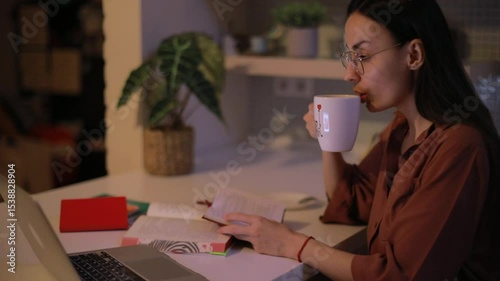 Young woman with long dark hair and glasses drinks from a mug while reading and working on a laptop at a cozy desk in the evening, surrounded by books and warm lighting.
