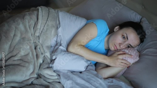 Young woman in a blue shirt lies in bed holding a soft pillow, looking  thoughtful, wrapped in a cozy blanket in a calm home setting.
