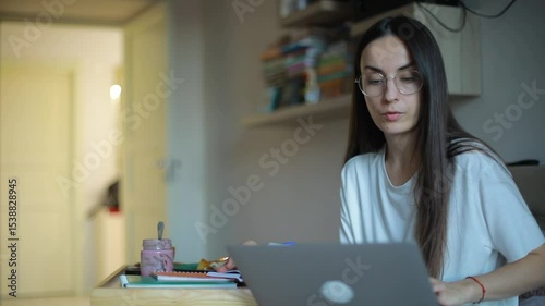 Young woman with long hair and glasses works on a laptop while sitting cross-legged on a bed in a cozy room, focused and relaxed in a casual home setting.
