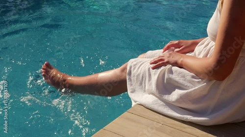 A woman in a white dress with wet legs is sitting on the edge of a wooden deck, dipping her feet into a swimming pool.