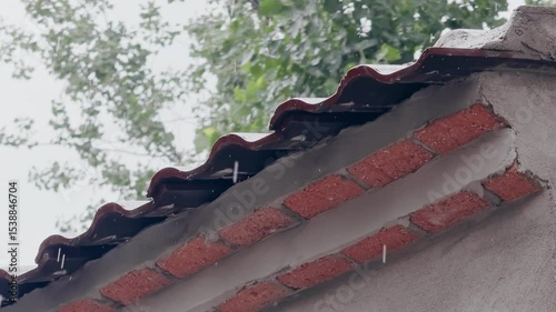 Raindrops falling from the red tile eaves of a rustic house during a rainy day, with lush green trees visible in the background.
