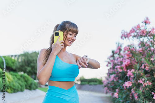 A smiling woman in activewear checks her fitness watch while holding a phone, enjoying an outdoor workout session in nature. Concept of fitness, tracking, and healthy living.