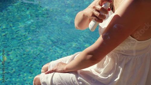 A woman in a white dress sprays sunscreen on her arm, with a bright blue swimming pool in the blurred background.
