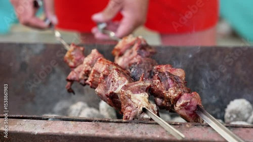 Hands adjust skewers of sizzling meat cooking over hot coals on a grill, highlighting the process of outdoor barbecue.