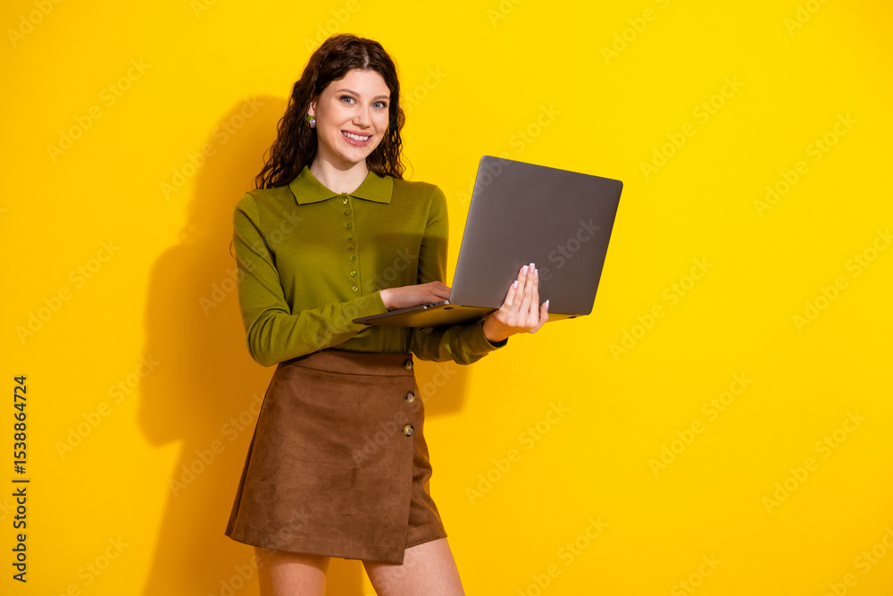 Fototapeta premium Cheerful young woman holding a laptop against a vibrant yellow background while showcasing modern casual style
