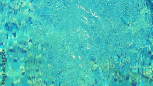 An overhead shot of a vibrant blue swimming pool, showing ripples and reflections on the water's surface, with mosaic tiles visible beneath.