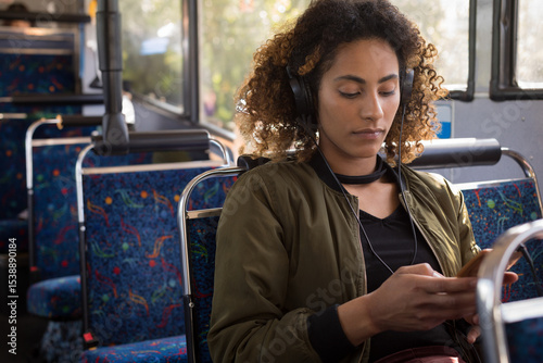 African American woman sitting on patterned bus seat wearing headphones scrolling smartphone photo