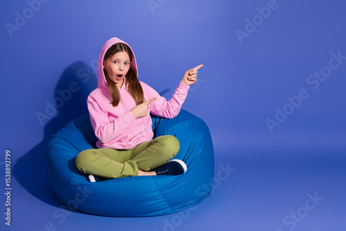 Preteen girl sitting on a blue bean bag chair pointing excitedly with her hands in a playful and causal hoodie outfit