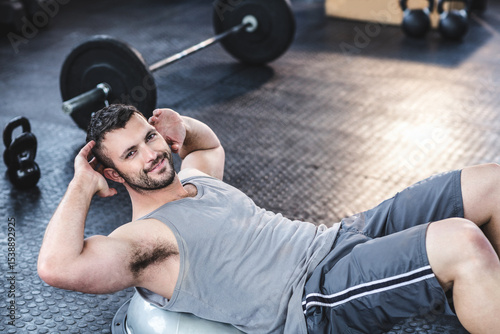 Male athlete doing crunches on hemispheric trainer beside barbell and kettlebells in gym