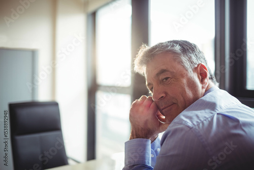 Man wearing blue dress shirt leaning at office desk next to chair and windows, copy space