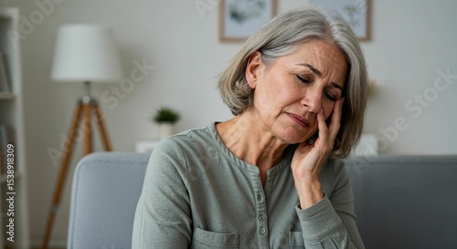 Senior caucasian woman experiencing ear pain at home, sitting on couch with hand on face and closed eyes, showing discomfort in indoor environment.
