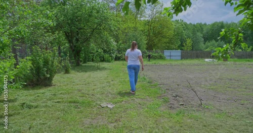 A woman in everyday clothes walking around a sunny garden, passing next to trees and a wooden fence with laid -back, calm behavior. The woman goes to get out in the garden.